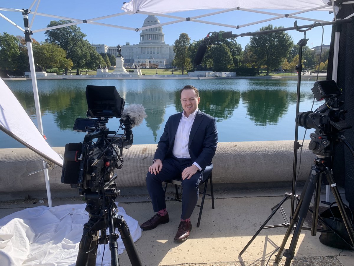 Seated for a broadcast interview beside the Capitol Reflecting Pool in Washington, cameras and lights around