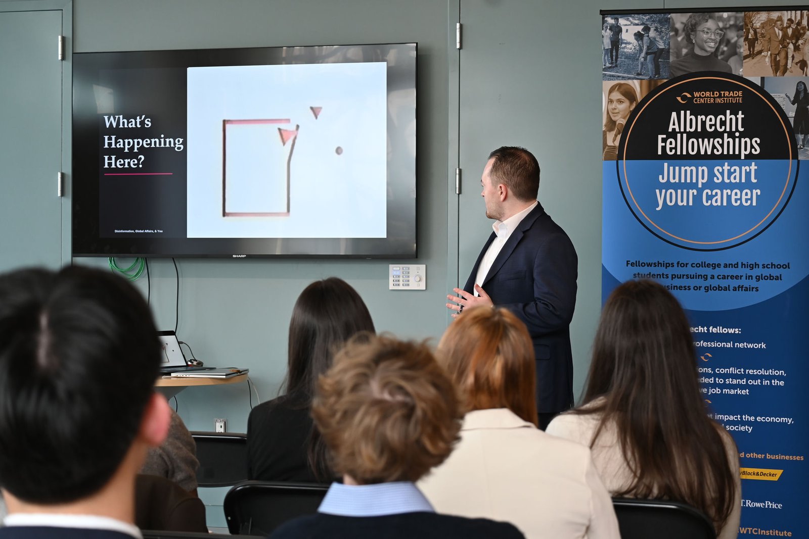 Presenting 'Disinformation, Global Affairs, & You' to a seated student audience beside a 'What's Happening Here?' slide, a World Trade Center Institute Albrecht Fellowships pull-up banner at right