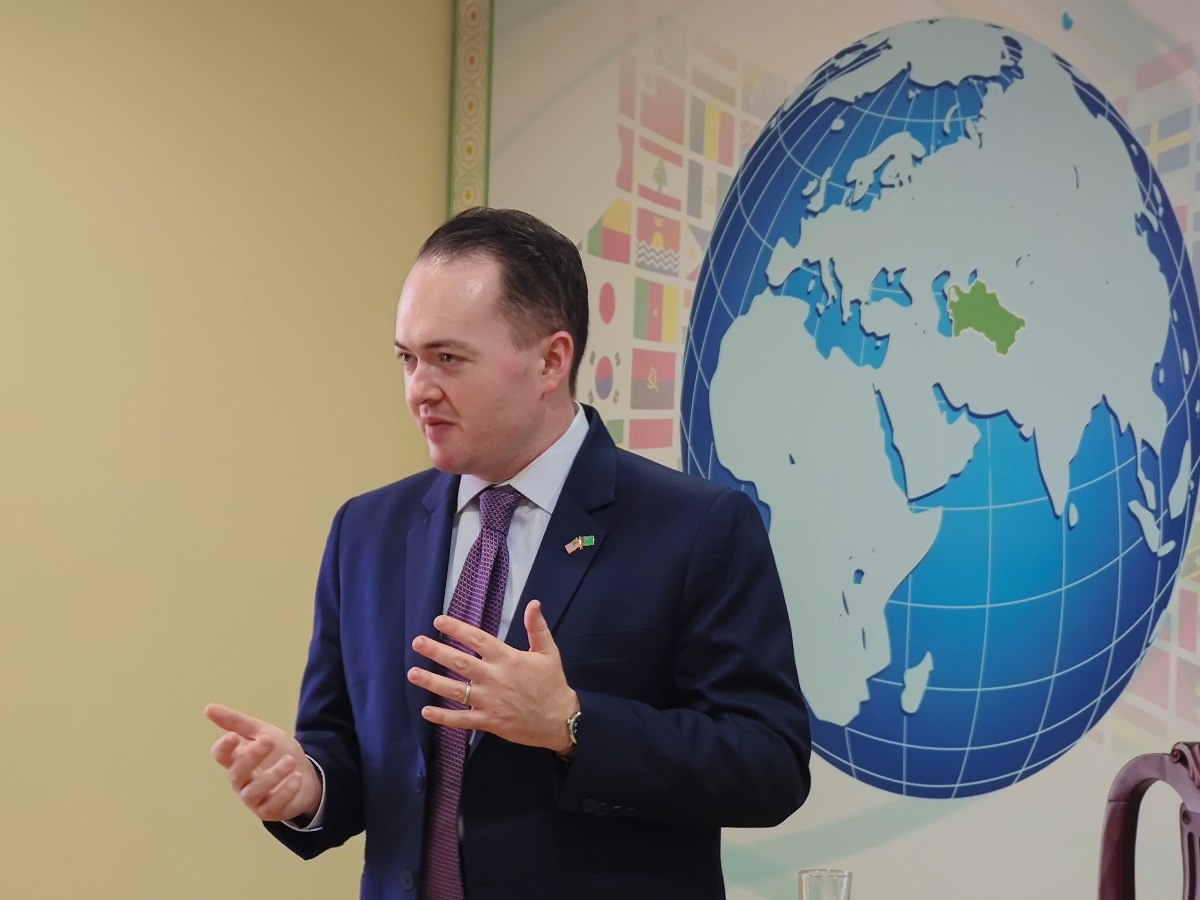 Speaking mid-gesture in front of a large painted globe mural ringed with the flags of many nations, an American and Turkmen flag pin on the lapel, Ashgabat