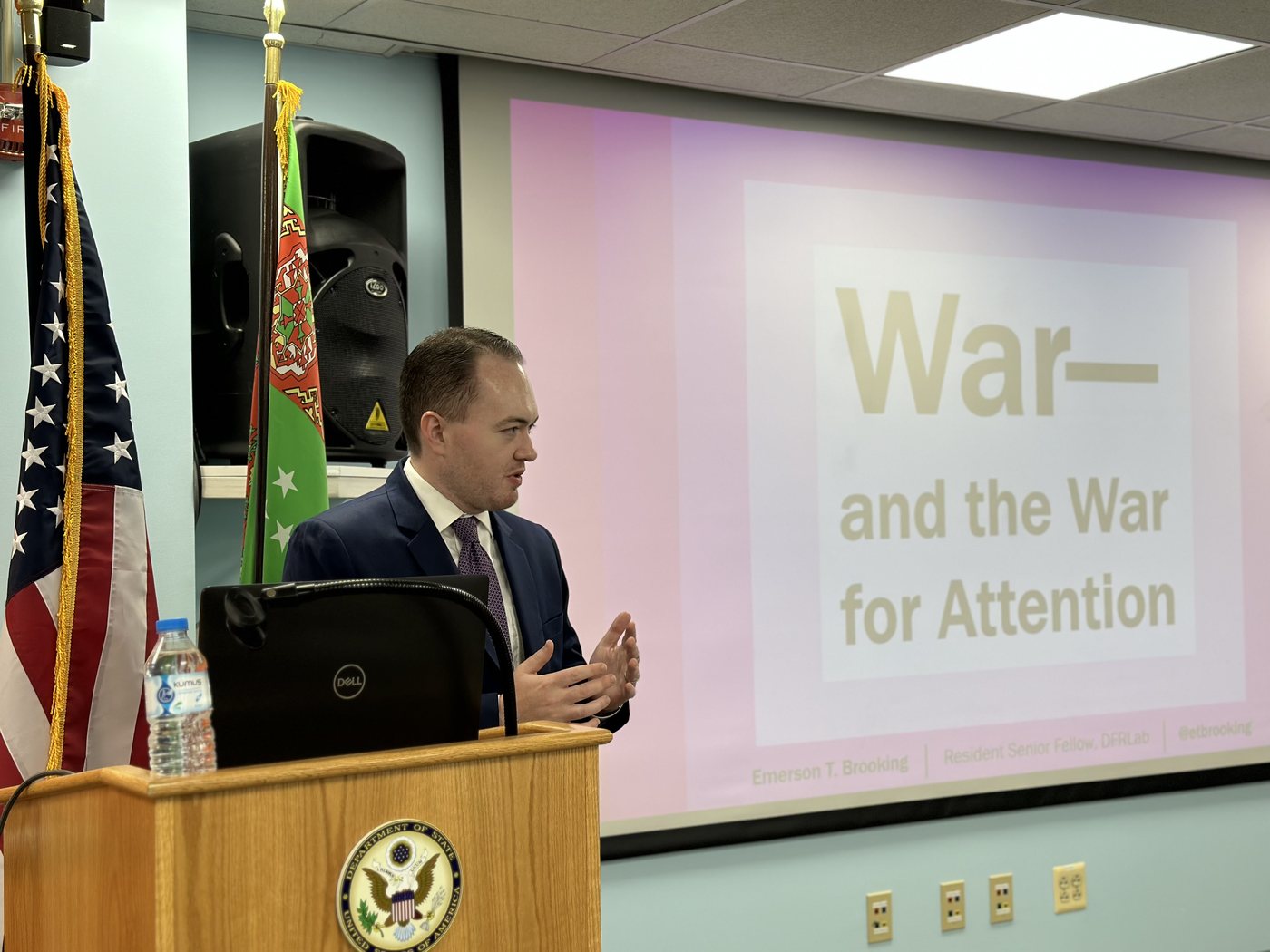 Speaking at a lectern beside American and Turkmen flags, projection slide reading War — and the War for Attention