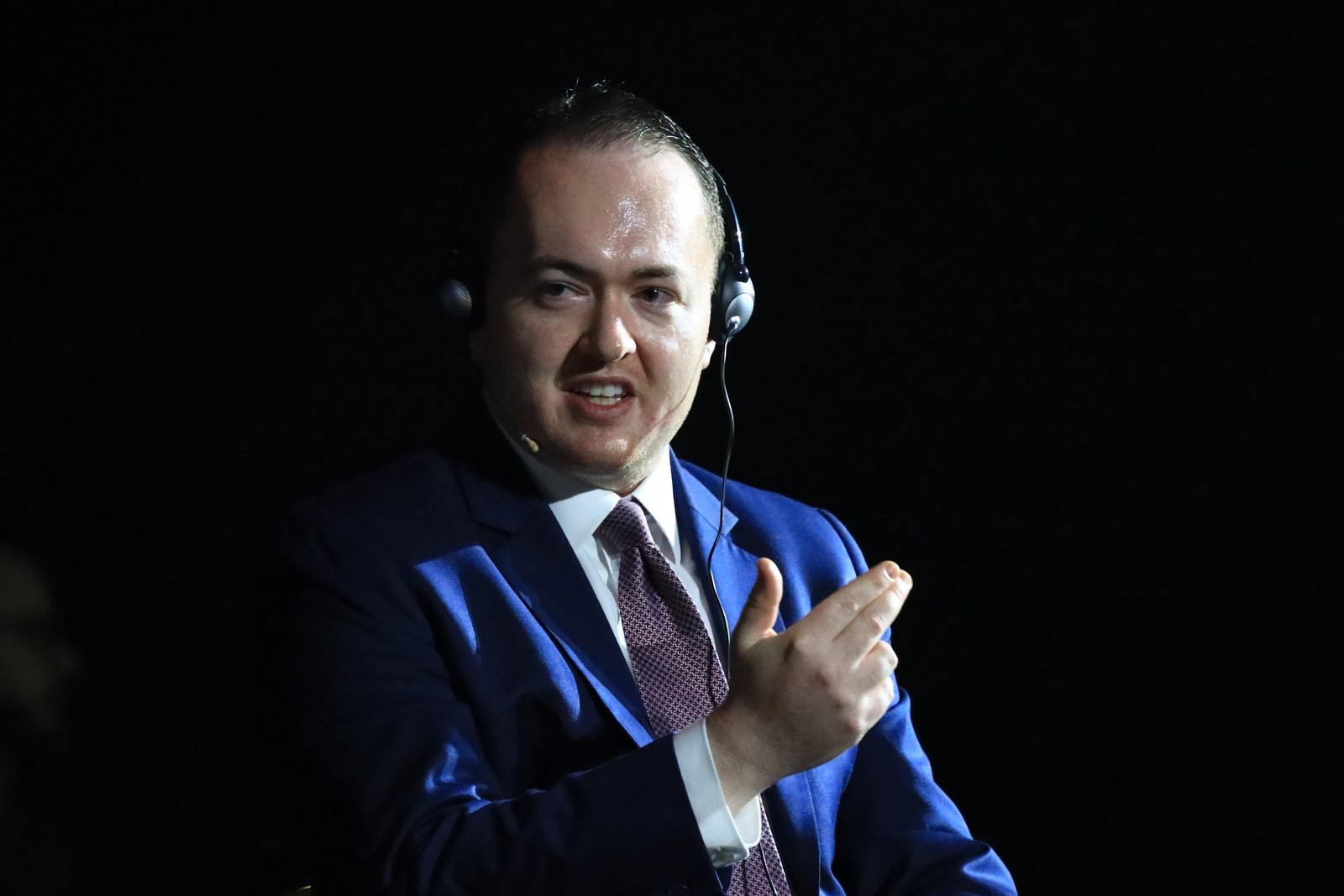 Tight close-up, mid-gesture, wearing an interpreter headset with a navy suit and patterned tie, lit by a single spotlight against black
