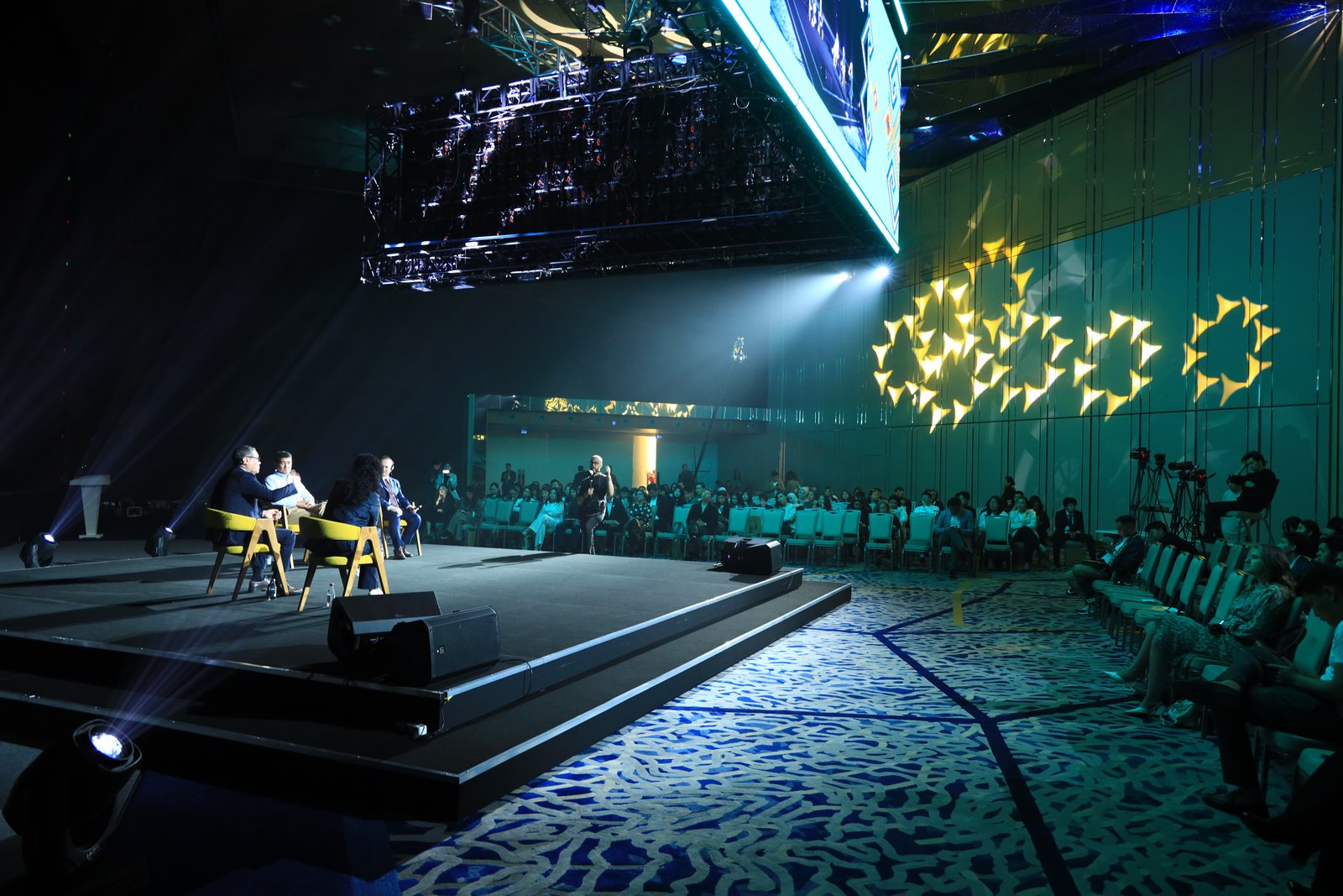 Wide view of the four-person QAZAQ Forum panel seated on a dark stage under a geometric LED backdrop, Astana, May 2024