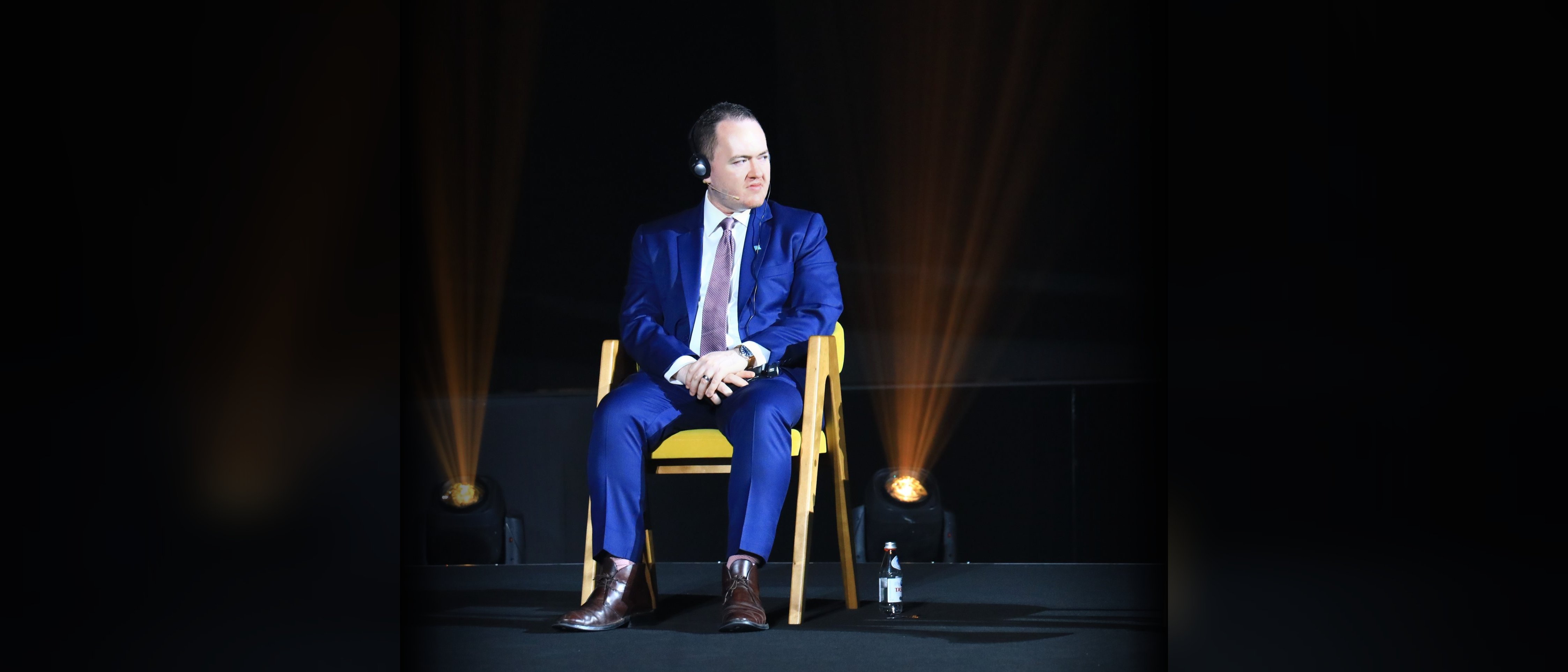 Seated on a dark theatrical stage under gold spotlight beams, in conversation with a host, wearing an interpreter headset