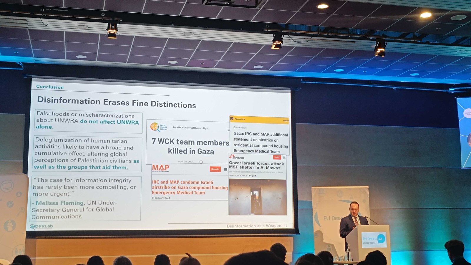 Seen past an audience's silhouetted heads, presenting at a lectern branded EU DisinfoLab, a large screen behind reading “Disinformation Erases Fine Distinctions” and quoting UN Under-Secretary-General Melissa Fleming, a DFRLab byline visible on the slide