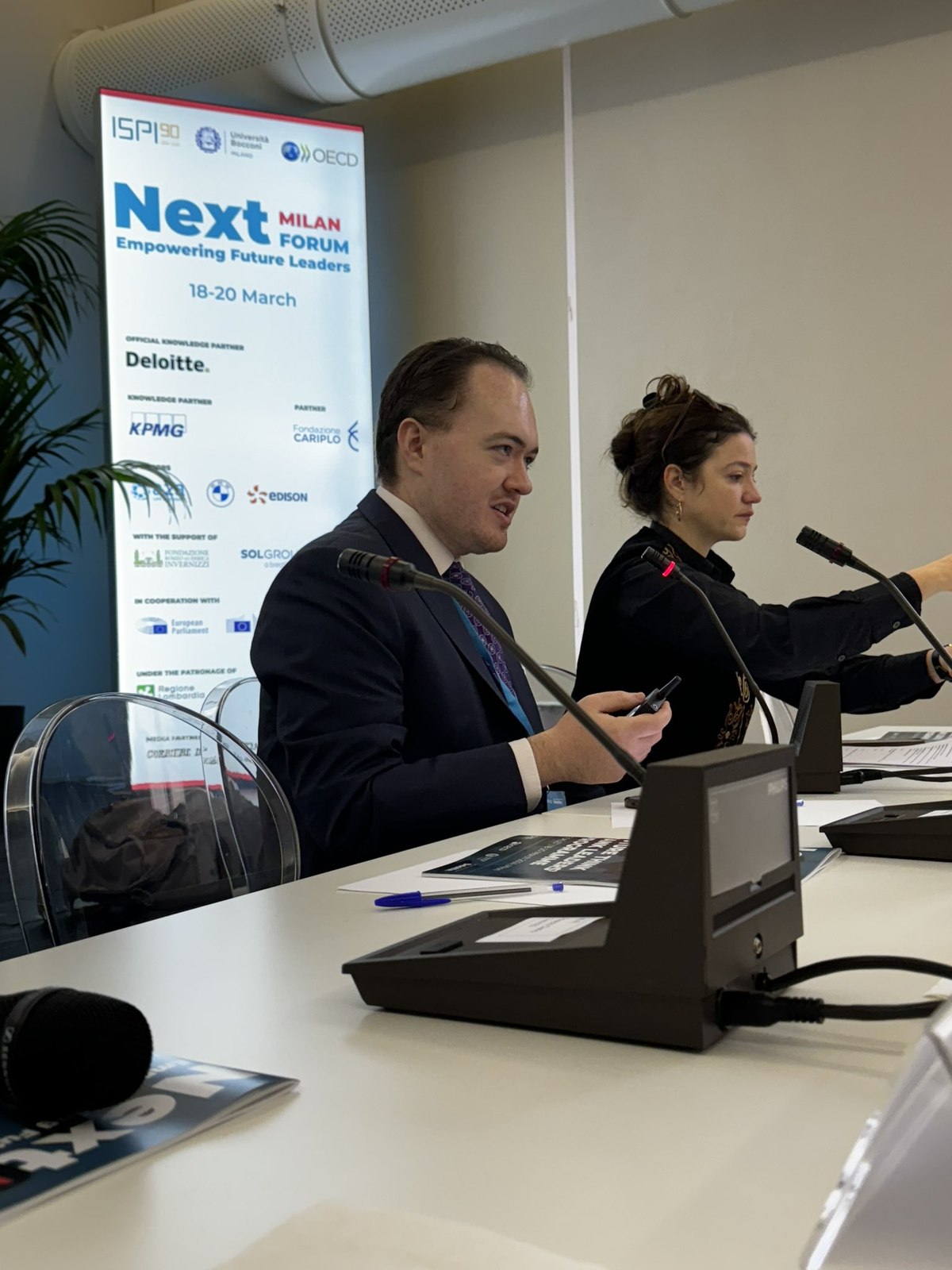 At a panel table with a microphone, gesturing with a pen mid-conversation, beside a co-panelist, in front of a “Next Milan Forum — Empowering Future Leaders, 18-20 March” step-and-repeat banner with ISPI, Bocconi, and OECD sponsor logos