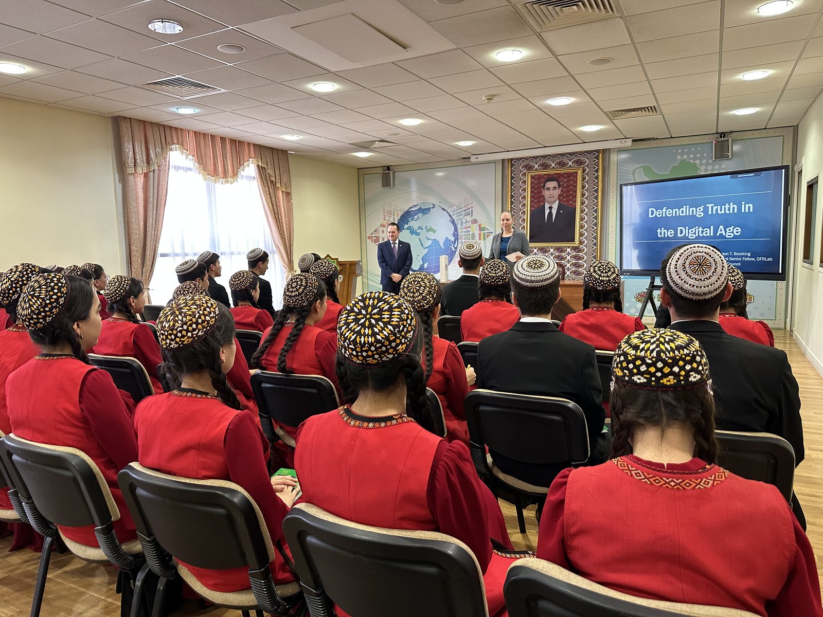 An auditorium of Turkmen students in traditional red dress and embroidered skullcaps, seen from behind, facing a screen that reads Defending Truth in the Digital Age