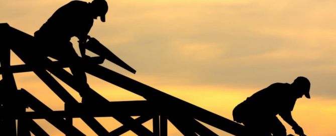 Silhouettes of two construction workers framing a roof against an orange sunset sky.