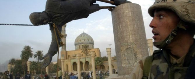 A U.S. soldier stands beside a toppled statue of Saddam Hussein in Baghdad, with a mosque in the background.