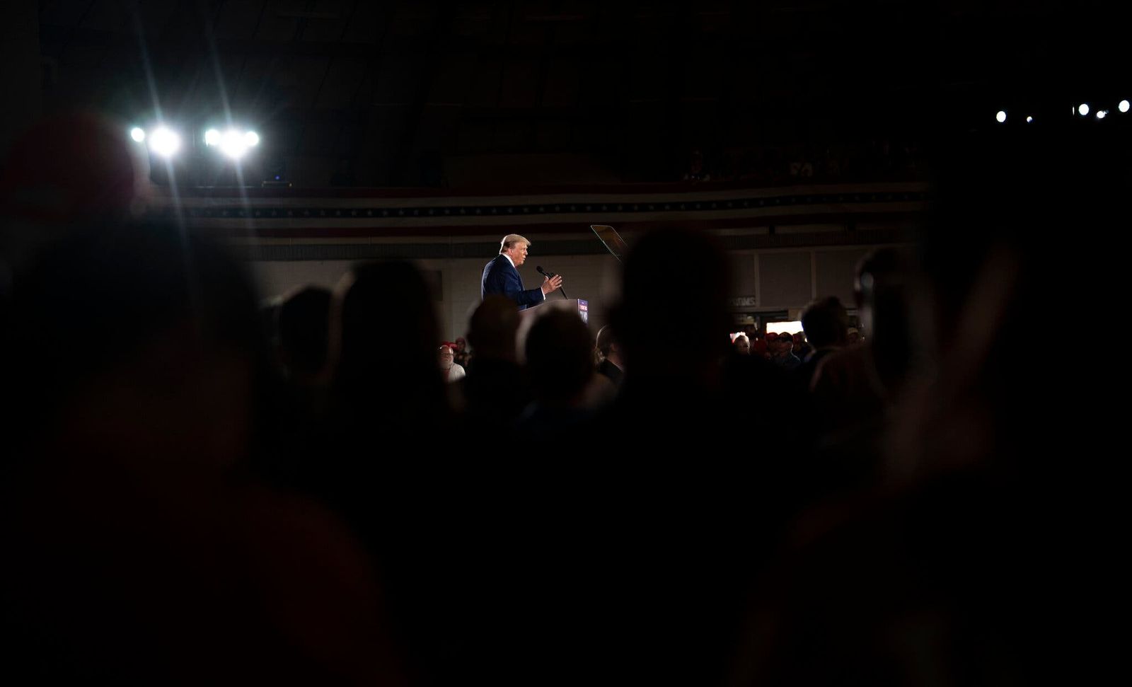 Donald Trump speaking at a podium on a dark stage, seen through a silhouetted crowd in a dim arena.